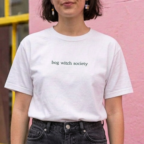 A woman wearing a white short-sleeve T-shirt with the words 'bog witch society'.