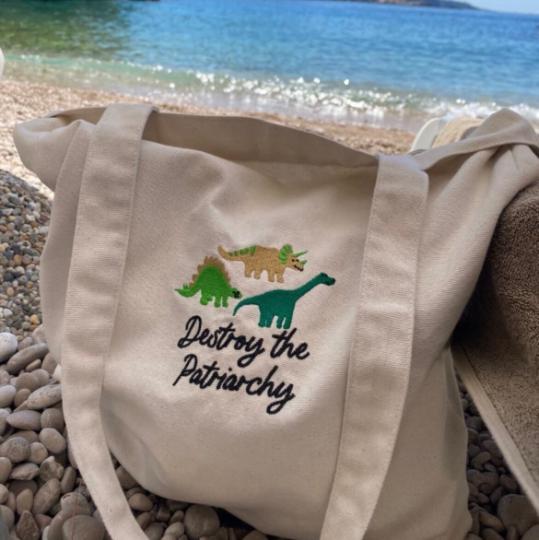 Destroy the Patriarchy embroidered dinosaur tote bag on a pebble beach