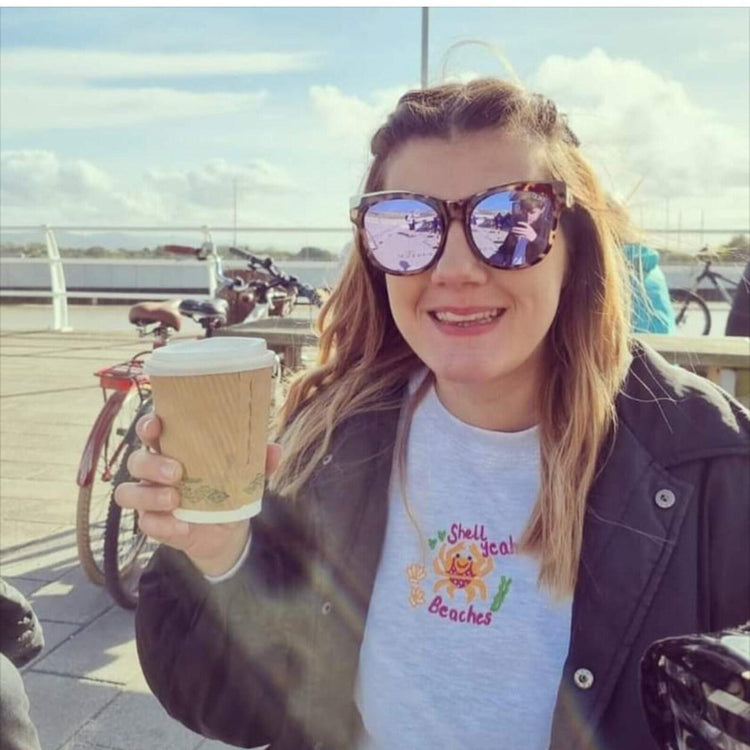 Woman holding a coffee wearing a grey Shell Yeah Beaches embroidered sweatshirt