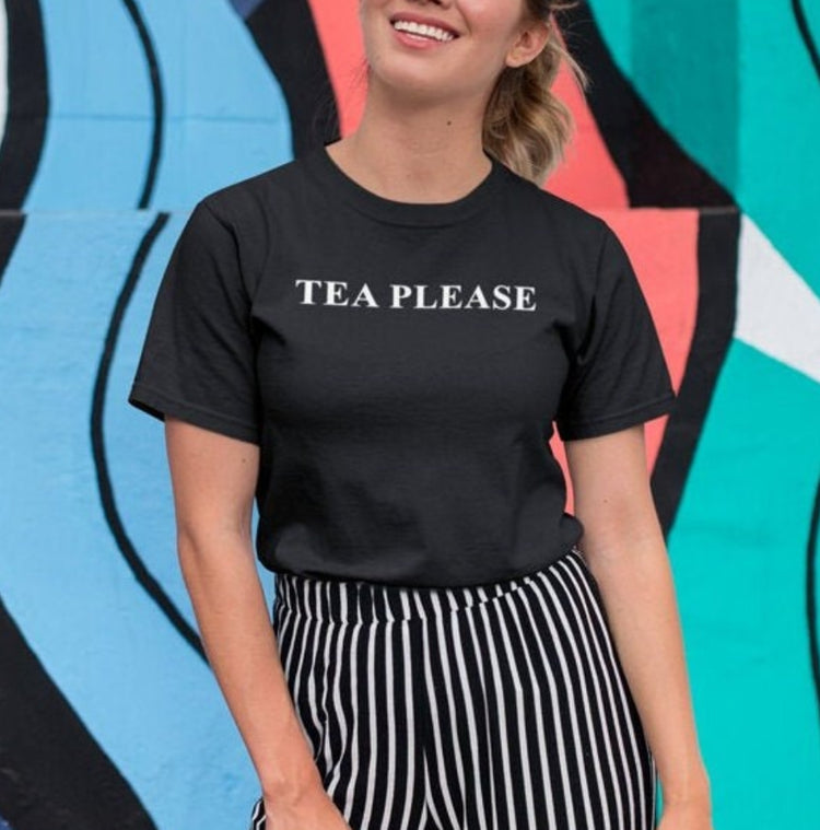 Woman in a black Tea Please tee in front of a colourful mural